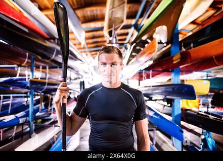 Portrat eines jungen Kanufahrers, der mitten in gestapelten Kanus steht. Das Konzept des Kanufahrens als dynamischer, abenteuerlicher Sport Stockfoto