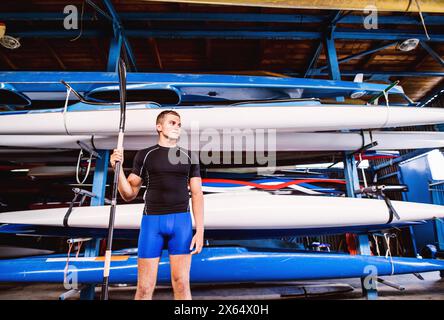 Portrat eines jungen Kanufahrers, der mitten in gestapelten Kanus steht. Das Konzept des Kanufahrens als dynamischer, abenteuerlicher Sport Stockfoto