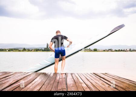Junger Kanufahrer, der Kanu ins Wasser bringt, auf Holzsteg steht. Das Konzept des Kanufahrens als dynamischer, abenteuerlicher Sport Stockfoto
