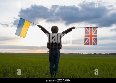 Das unerkennbare Kind hält britische und ukrainische Fahnen vor dem Sonnenuntergangshimmel. Flaggen der Ukraine und Großbritanniens. Konzept der Zusammenarbeit und des partn Stockfoto