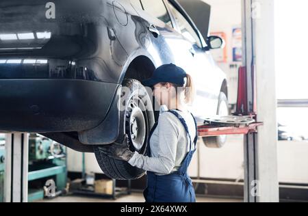 Weibliche Automechanikerin, die im Auto-Service die Stufen wechselt. Schöne Frau, die Reifen in einer Garage hält, blaue Overalls trägt. Reifenmontage. Weibliche Automobile Stockfoto