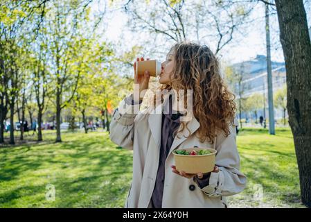 Junge Frau mit lockigen Haaren trinkt Kaffee und hält Salatschüssel, während sie in sonnigem Park steht, gesunder Lebensstil inmitten üppiger Vegetation. Büroangestellte verbringt ihre Mittagspause im Stadtpark. Stockfoto