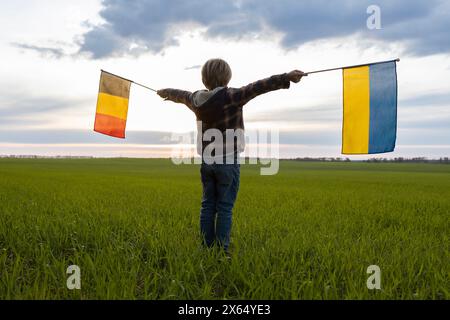 Unerkennbares Kind hält deutsche und ukrainische Fahnen vor dem Hintergrund des Sonnenuntergangs. Flaggen der Ukraine und Deutschlands. Konzept der Zusammenarbeit A Stockfoto