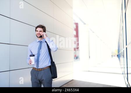 Gutaussehender Geschäftsmann telefoniert mit dem Smartphone, während er auf der Straße der Stadt läuft und ins Büro geht. Manager lächelt, im Freien in urbaner Umgebung Stockfoto