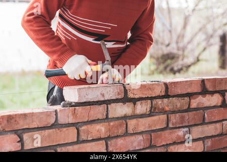 Ein Mann baut eine Ziegelmauer, legt einen Ziegel auf einen Zement-Sandmörtel und klopft mit einem Bauhammer auf den Ziegel Stockfoto