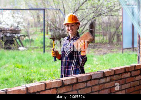 Eine junge Maurerin in einem orangen Bauhelm mit einem Bauhammer und einem Ziegel in den Händen steht und lächelt neben einer Ziegelmauer Stockfoto