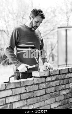 Ein Mann baut eine Ziegelmauer, legt einen Ziegel auf einen Zement-Sandmörtel, klopft mit einem Bauhammer auf den Ziegel Stockfoto