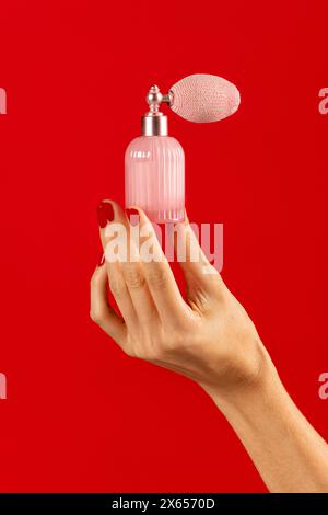 Anonyme junge weibliche dünne Finger mit nagellack, die glänzenden Kristall-Parfümbehälter mit Zerstäuber oben vor rotem Hintergrund innen halten Stockfoto