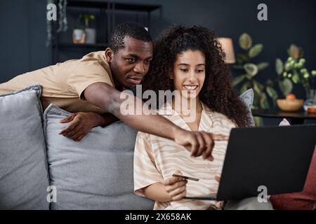 Mittlere Aufnahme eines jungen Schwarzen Mannes und einer birassischen Frau, die zu Hause Zeit zusammen verbringen, um online auf einem Laptop einzukaufen Stockfoto