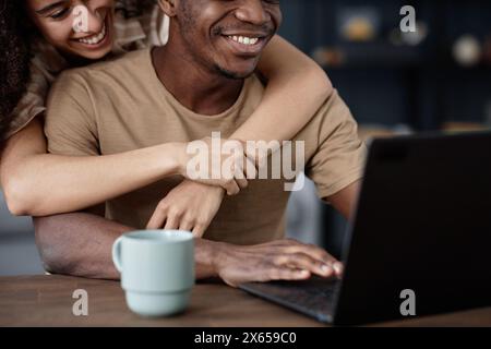 Eine fröhliche junge Frau, die ihren afroamerikanischen Freund umarmt, während sie auf einem Laptop im Internet surft Stockfoto