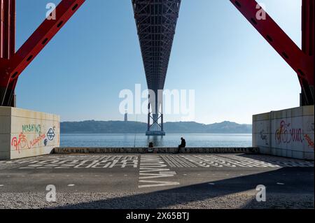 Unter der hoch aufragenden ponte 25 de abril erstreckt sich das Gebäude über den Fluss tejo; eine Einzelfigur ruht am Wasser auf einem mit Graffiti bedeckten Felsvorsprung. Stockfoto