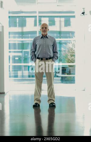 Ein reifer Geschäftsmann steht selbstbewusst in einer modernen Büroumgebung, sein blaues Hemd und seine khakifarbene Hose spiegeln ein professionelles Verhalten wider Stockfoto