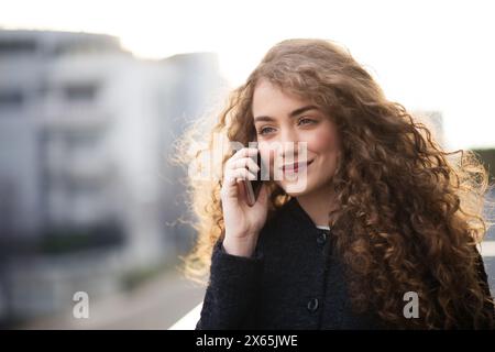 Wunderschöne Frau mit lockigem Haar, die auf der Terrasse stand und draußen telefonierte, während der kalten Frühlingstage. Morgensonne, Haare wehen im Wind. Stockfoto