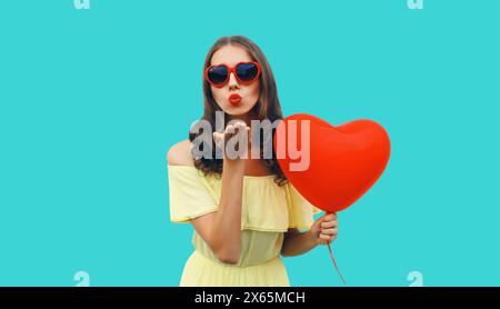 Porträt einer glücklichen jungen Frau mit rotem herzförmigem Ballon, der Kuss in der Sonnenbrille auf blauem Studiohintergrund bläst Stockfoto