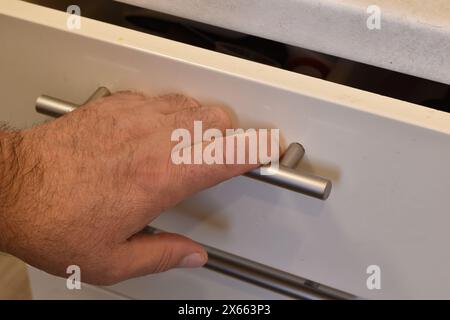 Das Foto zeigt die Hand eines Mannes, die auf dem Griff einer Schreibtischschublade liegt. Der Mann zieht die Schreibtischschublade heraus und greift den Metallgriff. Stockfoto