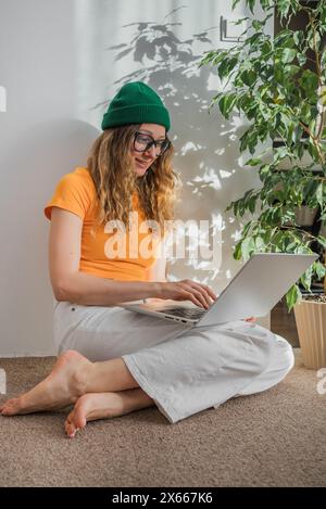 Die lächelnde junge Frau mit lockigem Haar, die eine Brille und eine grüne Mütze trägt, benutzt ihren Laptop bequem in einem sonnendurchfluteten Raum, umgeben von grünen Pflanzen. Stockfoto
