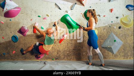 Kletterlehrer gibt einem Anfänger Unterricht auf einer Boulderwand in einem Fitnessstudio. Weibliche Athletin, die den Tablet-Computer hält und die richtigen Klettertechniken zeigt. Stockfoto