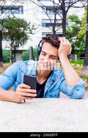 Der fröhliche junge Mann aus Denim lehnt sich an die Hand, während er Smartphone und Kopfhörer benutzt, in einem urbanen Park mit Bäumen und einem modernen Gebäude auf der Rückseite sitzt Stockfoto