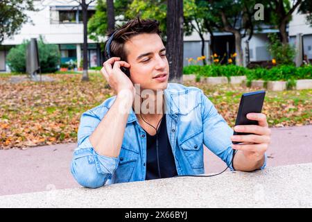 Ein junger Mann in einer Jeansjacke verwendet draußen Kopfhörer und ein Smartphone, lehnt sich entspannt und konzentriert auf eine Bank im Park. Stockfoto