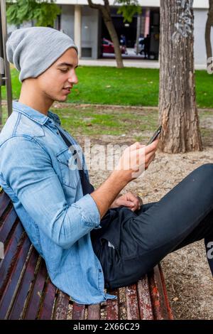 Ein fokussierter junger Mann in Jeansjacke und grauer Beanie sitzt auf einer Parkbank und nutzt sein Smartphone, umgeben von Bäumen und Gras. Stockfoto