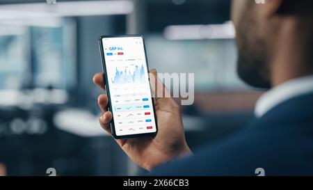 Schwarzer Geschäftsmann hält Smartphone und überprüft Aktien- und Kryptowährungsmarkt im Büro. Afroamerikanischer Geschäftsmann, der das Internet mit einem Mobiltelefon nutzt. Überschulteraufnahme Stockfoto