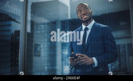 Porträt eines erfolgreichen schwarzen Geschäftsmannes im Anzug stehend mit Smartphone aus dem Fenster. Smiling Stock Market Investment Manager arbeitet am Abschluss des Geschäfts. Von außen aufgenommen Stockfoto