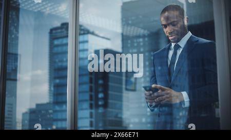 Porträt eines erfolgreichen schwarzen Geschäftsmannes im Anzug stehend mit Smartphone aus dem Fenster. Erfolgreicher CEO, der Investitionen in den E-Commerce plant. Stylische Außenaufnahme Stockfoto