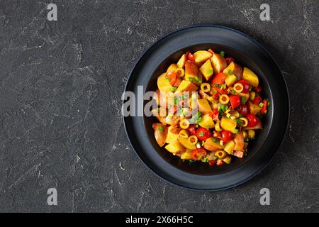 spanischer Kartoffelsalat aus Chorizo, grünen Oliven, Kirschtomaten, rotem Pfeffer und Koriander mit Honig-Senfdressing in schwarzer Schüssel auf Betontisch, Stockfoto