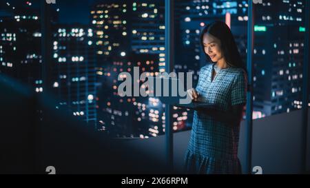 Finanzanalyst mit Laptop, neben dem Fenster stehend mit der wunderschönen Skyline der Nachtsstadt mit Wolkenkratzern. Selbstbewusste asiatische Geschäftsfrau, die in modernen Unternehmensbüros arbeitet. Stockfoto