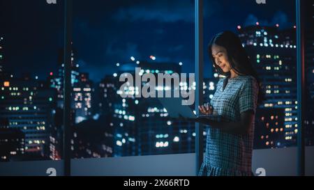 Finanzanalyst mit Laptop, neben dem Fenster stehend mit der wunderschönen Skyline der Nachtsstadt mit Wolkenkratzern. Selbstbewusste asiatische Geschäftsfrau, die in modernen Unternehmensbüros arbeitet. Stockfoto