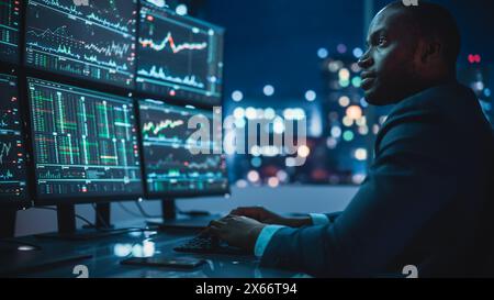 Finanzanalyst arbeitet am Computer mit Multi-Monitor Workstation mit Echtzeitbeständen, Rohstoffen und Börsendiagrammen. Ein afroamerikanischer Geschäftsmann arbeitet spät in der Nacht bei der Investment Bank. Stockfoto