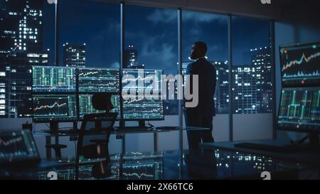 Erfolgreicher Geschäftsmann, der am späten Abend aus dem Fenster schaut. Modernes Hedge Fund Office mit Computer mit Multi-Monitor Workstation mit Echtzeit-Aktien, Rohstoffen und Börsenkarten. Stockfoto