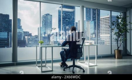 Erfolgreicher afroamerikanischer Geschäftsmann, der an seinem Schreibtisch sitzt und in seinem Büro in Big City an einem Laptop arbeitet. Handsome Corporation CEO Planning Investment Strategy for e-Commerce Project. Stockfoto