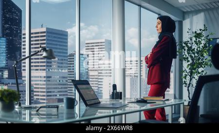 Erfolgreiche muslimische Geschäftsfrau im Anzug und Burka steht im Büro und blickt aus dem Fenster auf Big City. Selbstbewusste digitale Unternehmerinnen planen Investitionsstrategie für E-Commerce-Startup Stockfoto