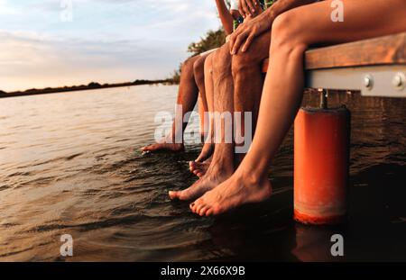 Vier junge Freunde sitzen an einem Sommertag auf dem Pier mit ihren Beinen am Wasser. Stockfoto