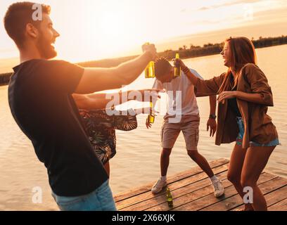 Gruppe junger Freunde, die Spaß beim Trinken von Bier und Tanzen auf dem Pier am See haben. Stockfoto