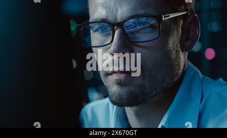 Bei Nacht Porträt eines gutaussehenden Startup Digital Entrepreneur, der am Computer arbeitet, Codezeile reflektiert in Brillen. Entwickler arbeiten an einer innovativen E-Commerce-App mit KI-Algorithmus Big Data. Blau Stockfoto