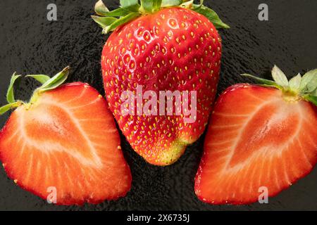 Nahaufnahme einer Erdbeere und zwei halbe Erdbeeren links und rechts auf schwarzem Hintergrund. Erdbeeren mit grünen Blättern. Stockfoto
