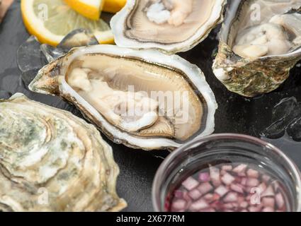 Geöffnete rohe Austern mit Soße und Zitronenscheiben auf grauem Steinservierbrett. Delikatessen. Stockfoto