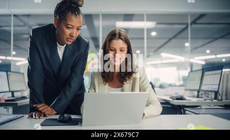Zwei weibliche Kollegen diskutieren liebevoll über die Arbeit, lächeln, während sie Project on Laptop in diversen modernen Geschäftsbüros zeigen. Erfahrener Manager und junger Mitarbeiter diskutieren analytische Daten. Stockfoto