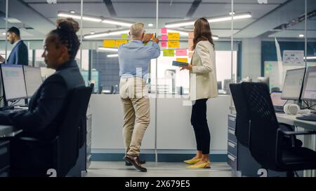 Junger Manager erstellt Projektplan mit einem erfahrenen Senior-Mitarbeiter auf der Bürowand mit Notizen in Papierform. Kollegen diskutieren Geschäfts-, Finanz- und Marketingprojekte. Spezialisten arbeiten in diversen Teams. Stockfoto
