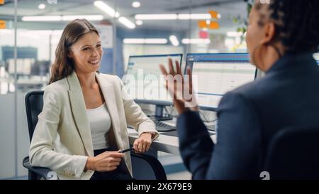 Zwei weibliche Kollegen sprechen liebevoll miteinander, lachen und lächeln, während sie an Computern in verschiedenen modernen Geschäftsbüros arbeiten. Erfahrener Manager und junger Mitarbeiter diskutieren ein unterhaltsames analytisches Projekt. Stockfoto