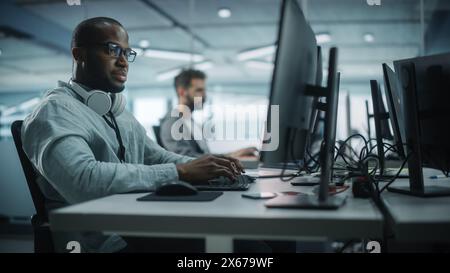 Diverse Büros: Black IT Programmierer arbeitet auf Desktop-Computern. Männlicher Spezialist, Der Innovative Software Engineer Entwickelt App, Programm, Videospiel. Terminal mit Codierungssprache. Stockfoto
