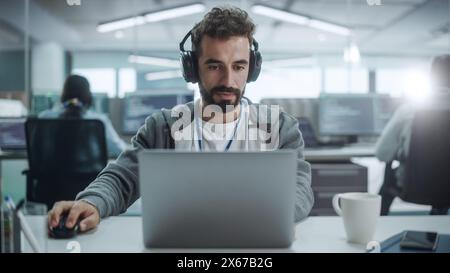 Büro: Porträt eines lächelnden IT-Programmierers, der Kopfhörer trägt, der auf einem Desktop-Computer arbeitet. Männlicher Websiteentwickler, Software Engineer, Der App Entwickelt, Videospiel. Podcast hören, Musik. Vorderansicht Stockfoto