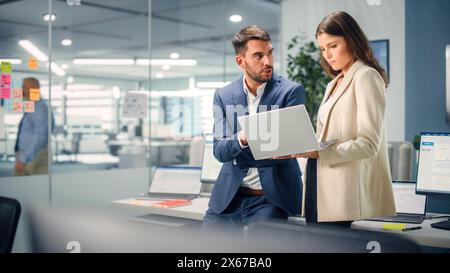 Junger Manager, der mit einem erfahrenen Kollegen spricht, während er im Büro einen Laptop verwendet. Kollegen diskutieren Geschäfts-, Finanz- und Marketingprojekte. Spezialisten arbeiten in diversen Teams. Stockfoto