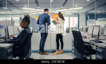 Junger Manager erstellt Projektplan mit einem erfahrenen Kollegen auf der Bürowand mit Notizen in Papierform. Mitarbeiter diskutieren Geschäfts-, Finanz- und Marketingprojekte. Spezialisten arbeiten in diversen Teams. Stockfoto