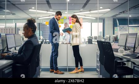 Junger Manager erstellt Projektplan mit einem erfahrenen Kollegen auf der Bürowand mit Notizen in Papierform. Mitarbeiter diskutieren Geschäfts-, Finanz- und Marketingprojekte. Spezialisten arbeiten in diversen Teams. Stockfoto