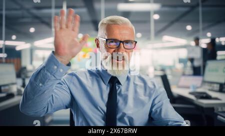 Porträt eines gutaussehenden erfahrenen Senior Managers, der einem Kunden bei Videoanrufen in Modern Office zuwinkt. Geschäftskonferenz mit Partnern, Kollegen, Familienmitgliedern oder Freunden am Computer. POV-Ansicht. Stockfoto
