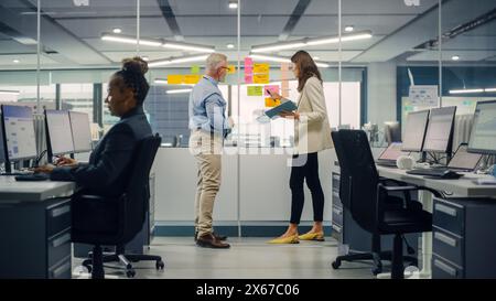 Junger Manager erstellt Projektplan mit einem erfahrenen Senior-Mitarbeiter auf der Bürowand mit Notizen in Papierform. Kollegen diskutieren Geschäfts-, Finanz- und Marketingprojekte. Spezialisten arbeiten in diversen Teams. Stockfoto