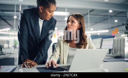 Zwei weibliche Kollegen diskutieren liebevoll über die Arbeit, lächeln, während sie Project on Laptop in diversen modernen Geschäftsbüros zeigen. Erfahrener Manager und junger Mitarbeiter diskutieren analytische Daten. Stockfoto